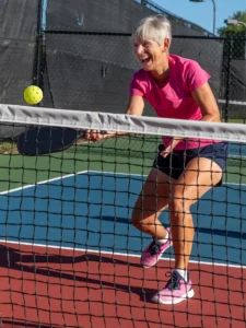 Smiling senior woman in a pink shirt reaching over the net for a pickleball volley on an outdoor court