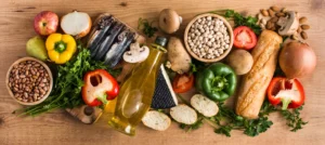 Mediterranean diet foods including olive oil, sardines, chickpeas, beans, almonds, fresh vegetables and crusty bread on a wooden table