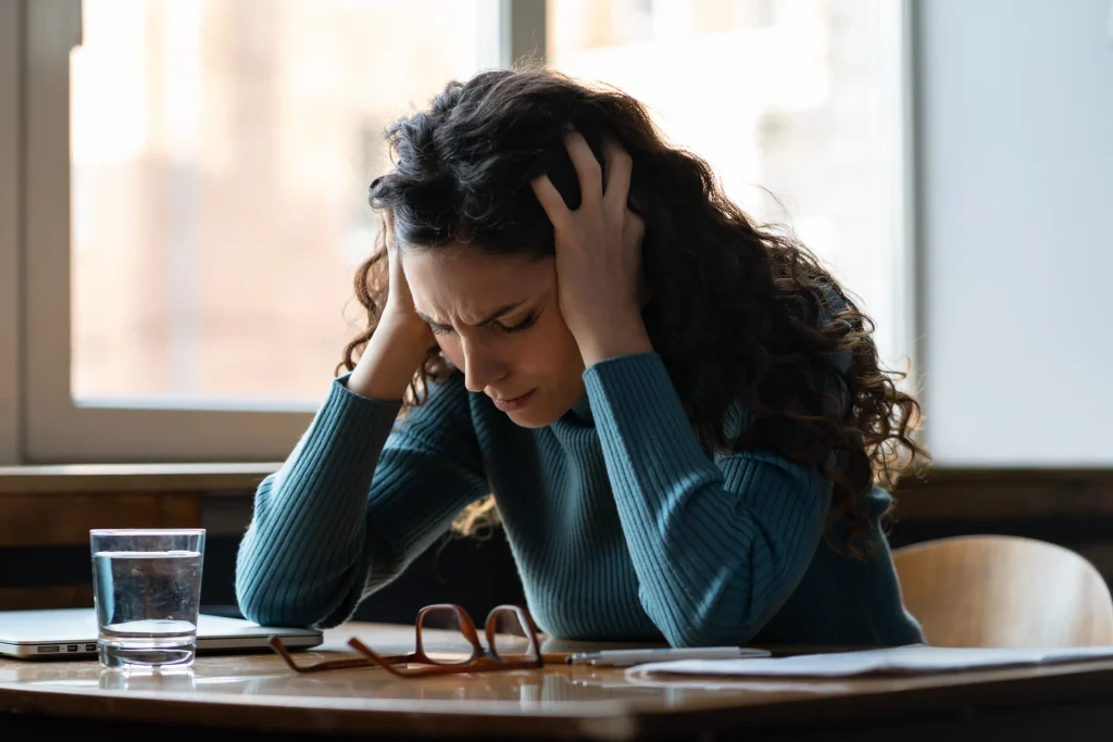 Woman at a desk holding her head in discomfort, illustrating how stress and lifestyle and inflammation Central Coast clients experience can show up as headaches and fatigue